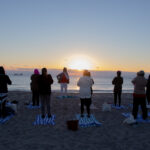 View of yoga students standing on the beach facing the sun and water